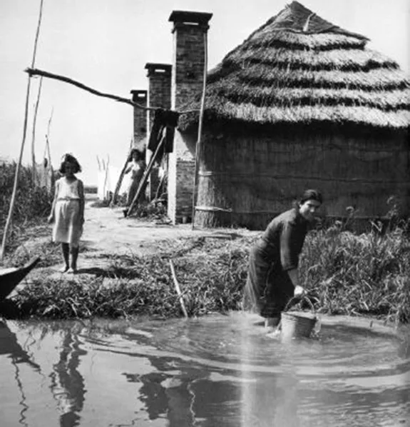 Donna che attinge acqua da un canale davanti a un casone con tetto in canna, fotografia storica